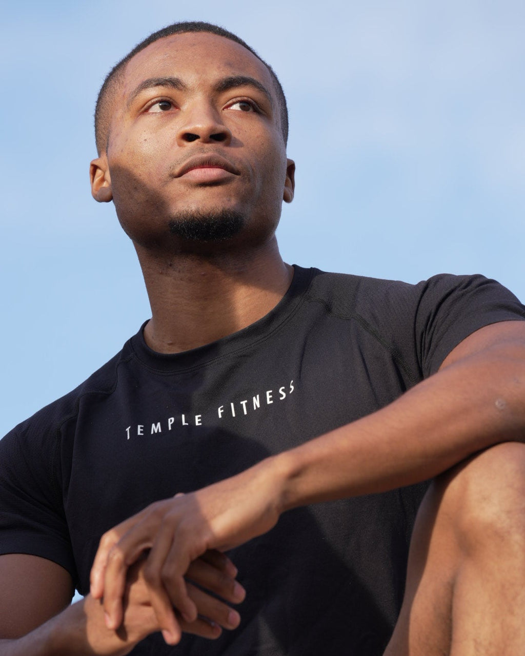Man wearing a black 'Temple Fitness' t-shirt against a clear blue sky